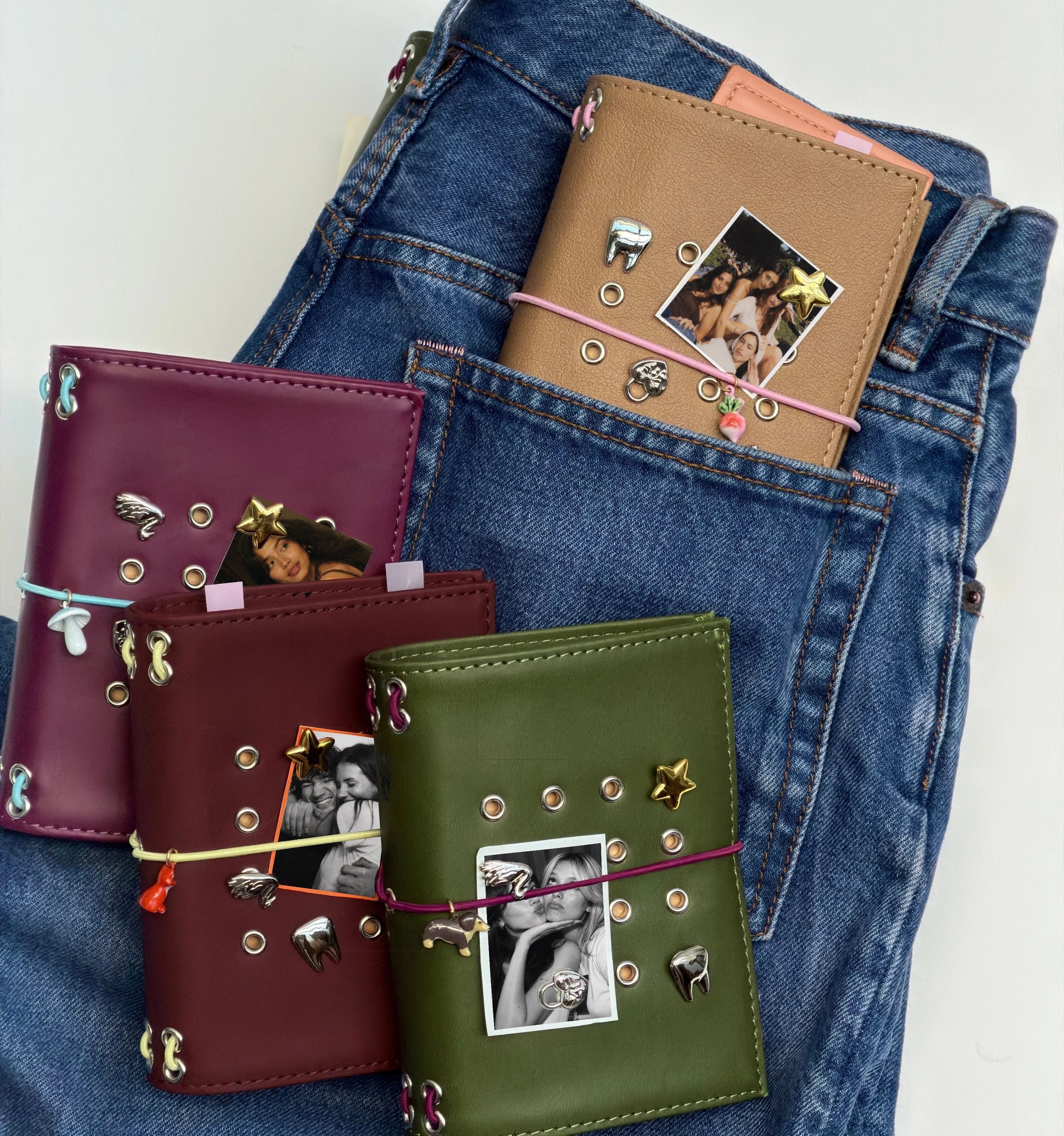 Four pocket journals in burgundy, camel, chestnut fox, and olive styled with removable metal charms, elastic bands, and personal photos, arranged on folded denim.