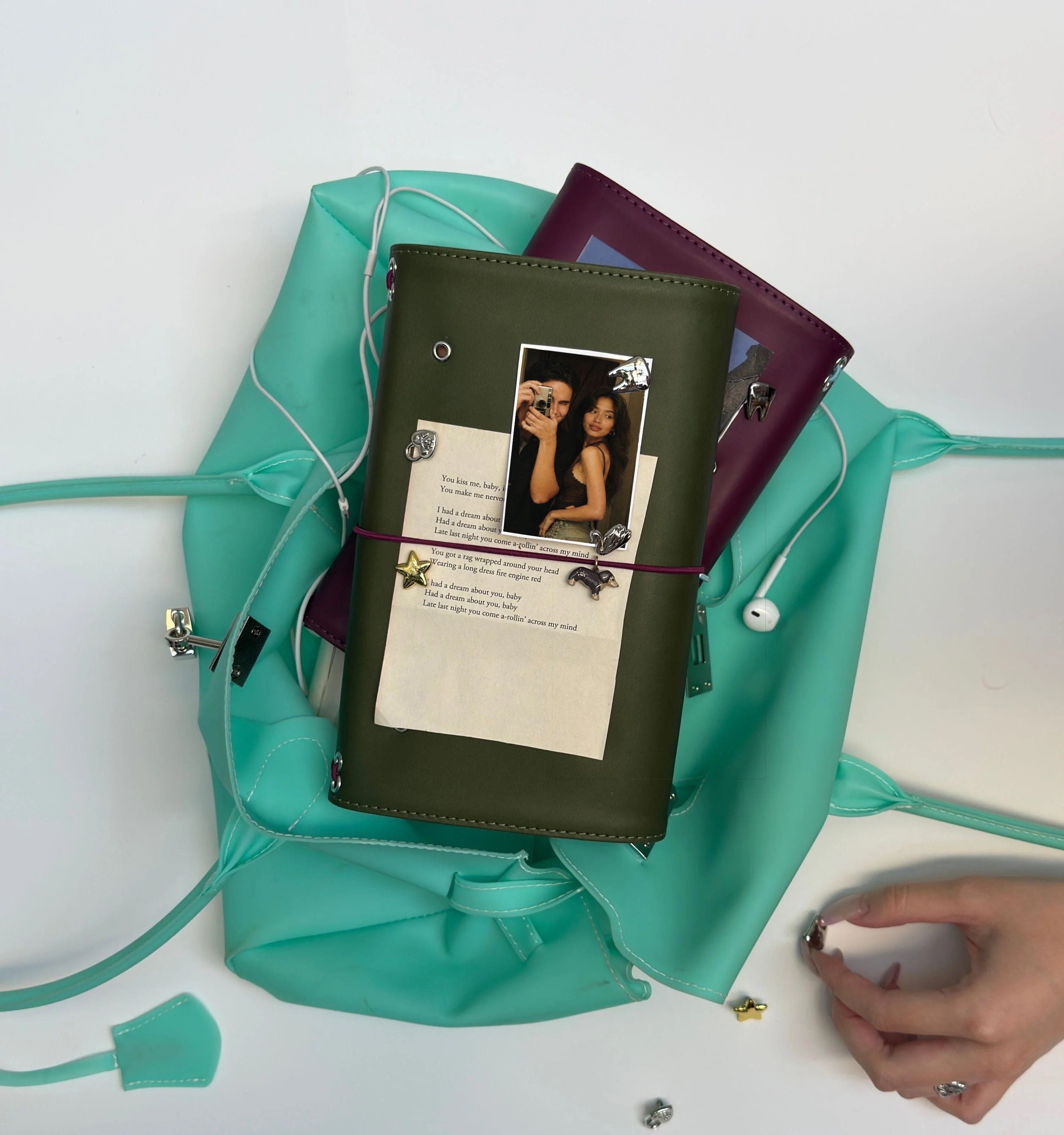 Olive and burgundy full-size journals styled with removable metal charms, personal photos, and paper keepsakes, arranged on a turquoise handbag.