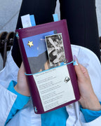 A person holds a Burgundy Full Size Journal decorated with a powder-blue elastic band, blue mushroom charm, removable KEPT cover charms, a portrait photo, and paper keepsakes.