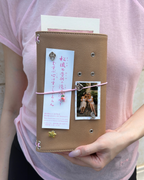 A person holds a Camel Radish Full Size Journal styled with a blush-pink elastic band, pink radish charm, four removable KEPT cover charms, a photo, and paper keepsakes.