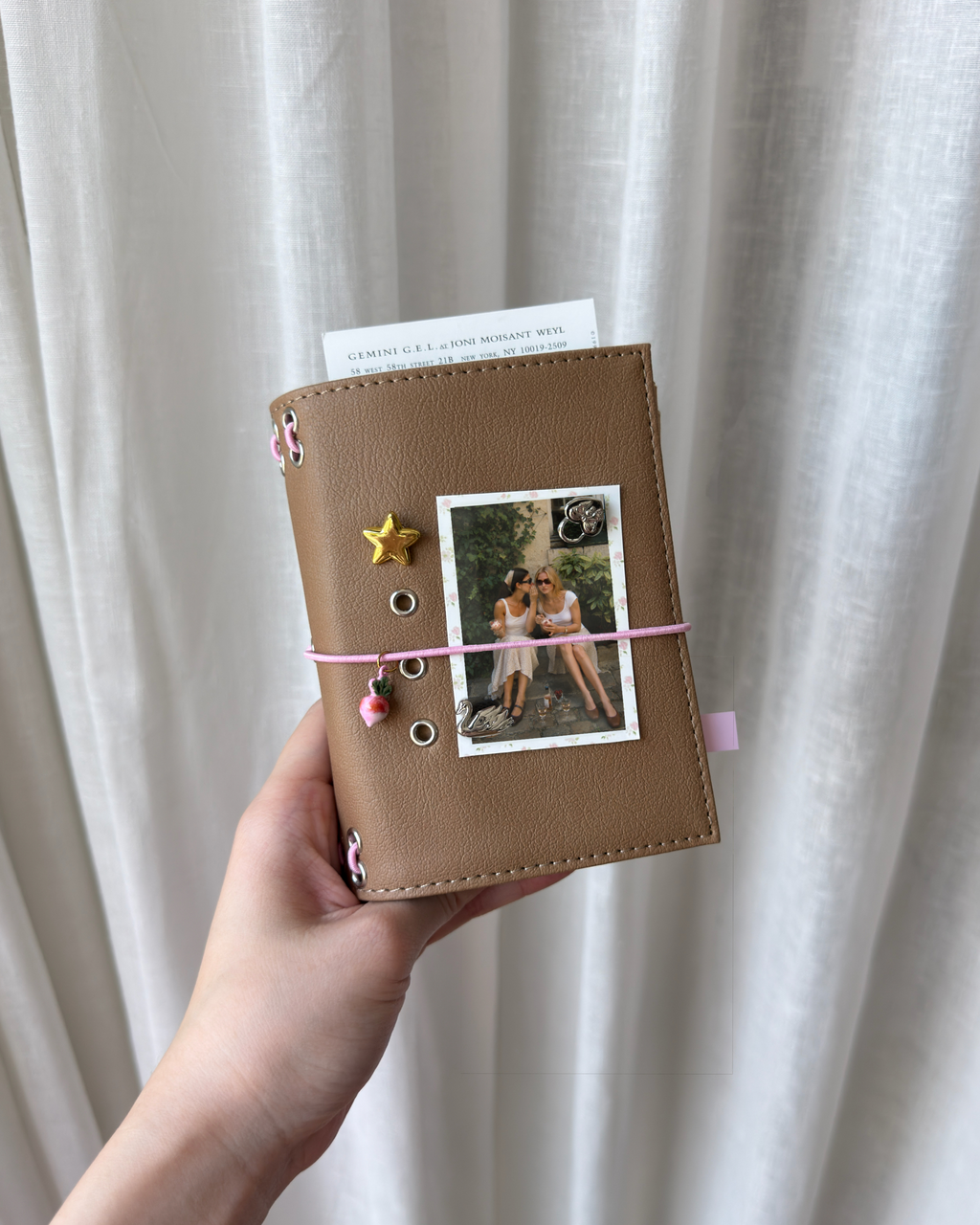 A hand holds a Camel Radish Pocket Journal styled with a blush-pink elastic band, pink radish band charm, removable KEPT cover charms, and a photo insert.