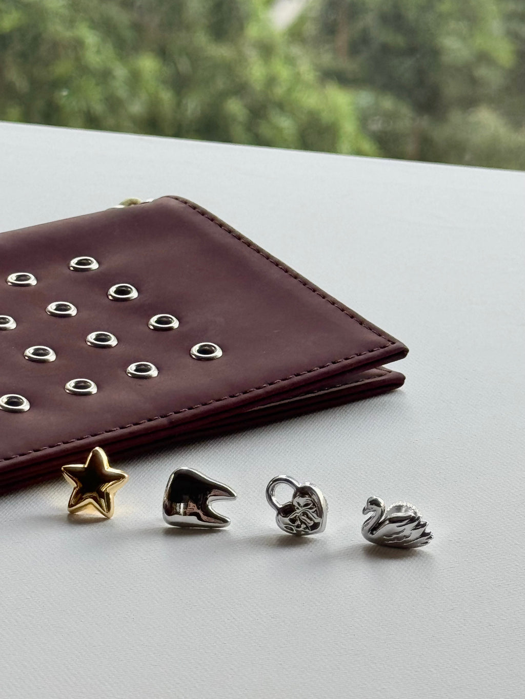 Four small jewelry charms or beads in gold and silver tones arranged on a light surface next to a burgundy leather wallet with silver studs, with a blurred green outdoor background.