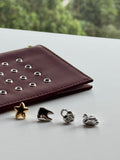Four small jewelry charms or beads in gold and silver tones arranged on a light surface next to a burgundy leather wallet with silver studs, with a blurred green outdoor background.