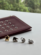 Four small jewelry charms or beads in gold and silver tones arranged on a light surface next to a burgundy leather wallet with silver studs, with a blurred green outdoor background.