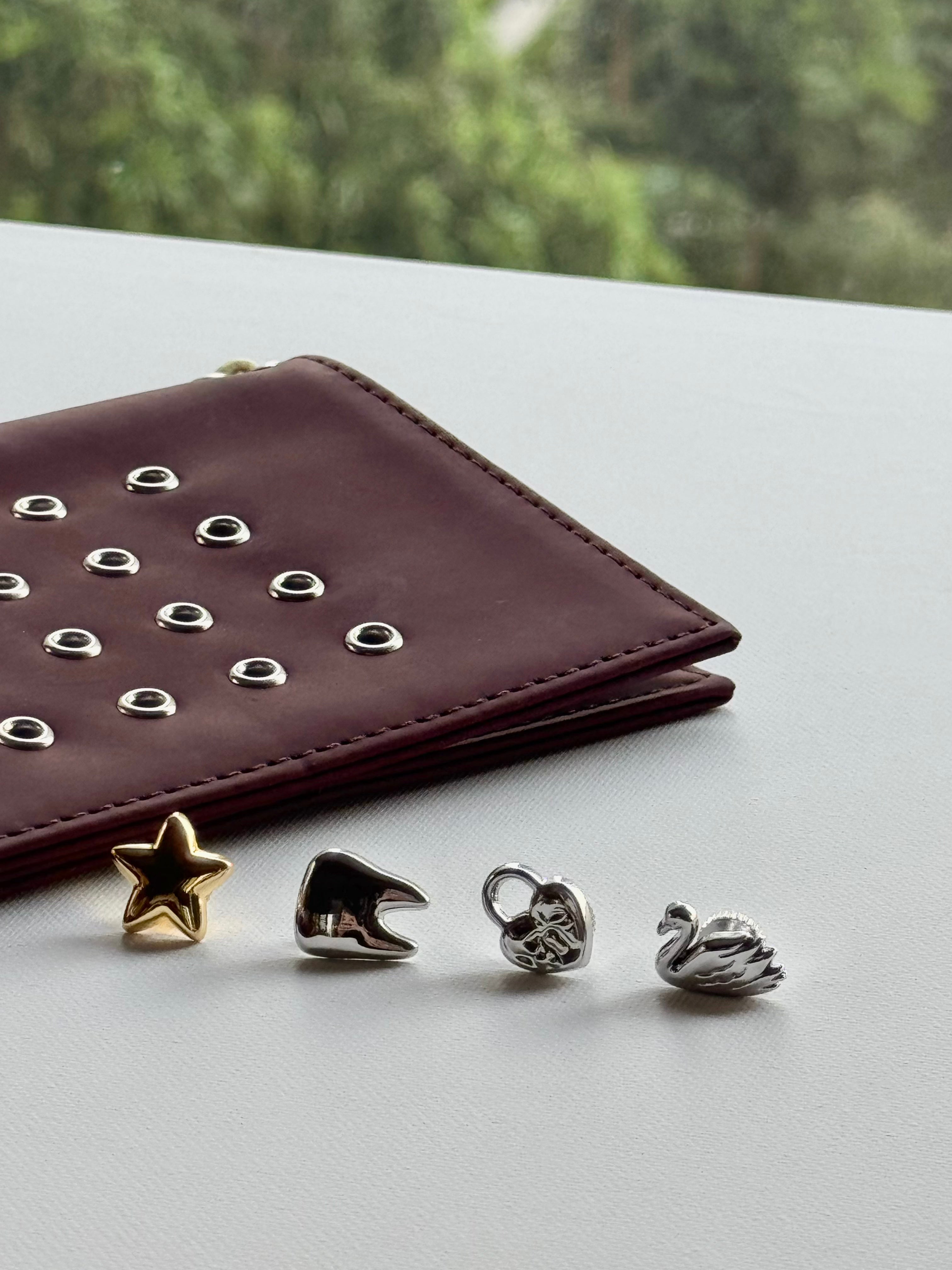 Four small jewelry charms or beads in gold and silver tones arranged on a light surface next to a burgundy leather wallet with silver studs, with a blurred green outdoor background.