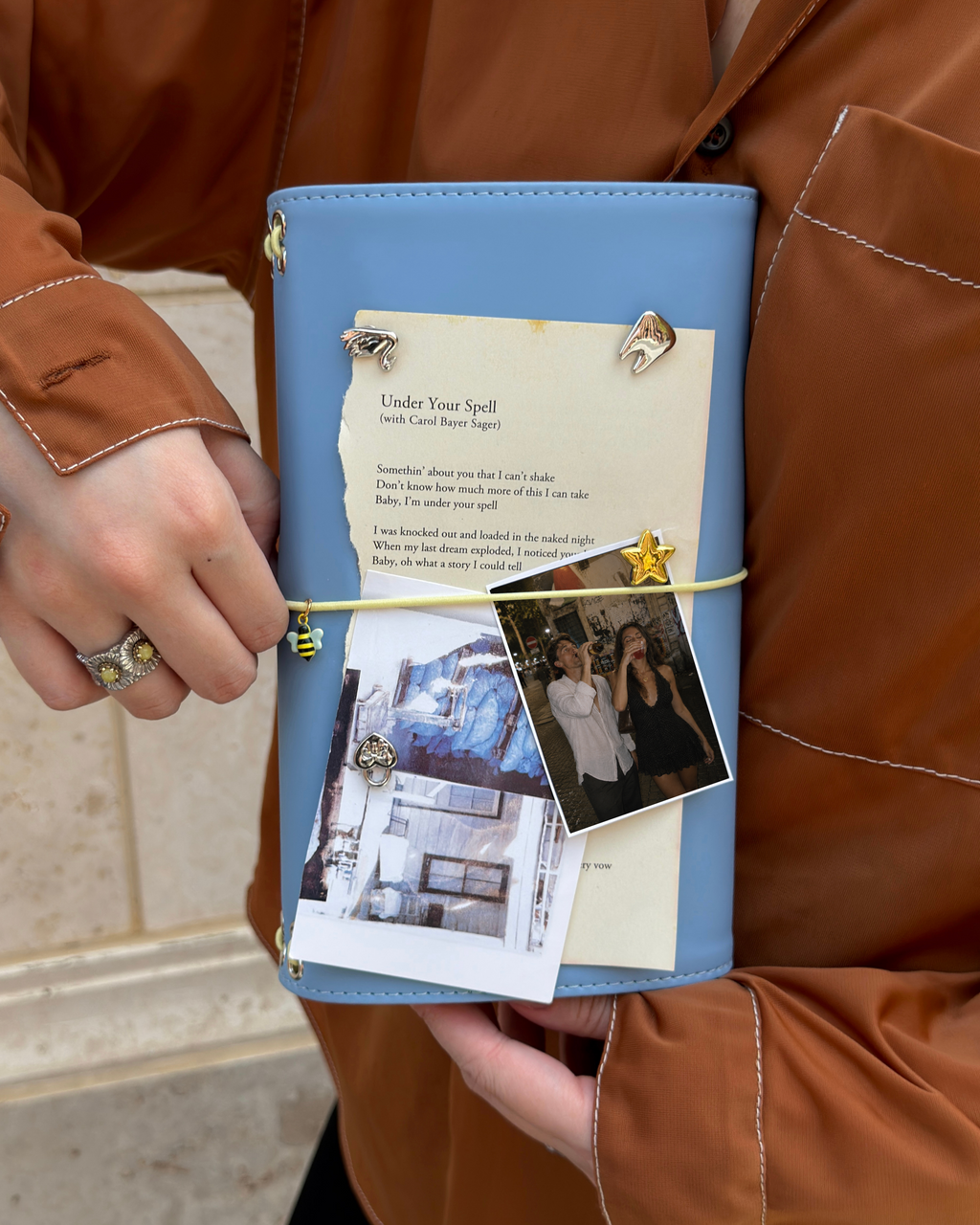 A person holds a Mist Blue Full Size Journal styled with a butter-yellow elastic band, bee charm, removable KEPT cover charms, a photo, and layered paper keepsakes.