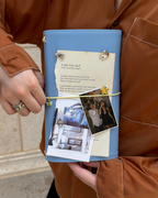 A person holds a Mist Blue Full Size Journal styled with a butter-yellow elastic band, bee charm, removable KEPT cover charms, a photo, and layered paper keepsakes.