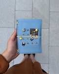 A hand holds a Mist Blue journal styled with a butter-yellow elastic band, gold eyelets, four removable KEPT charms, and a black-and-white photo insert.
