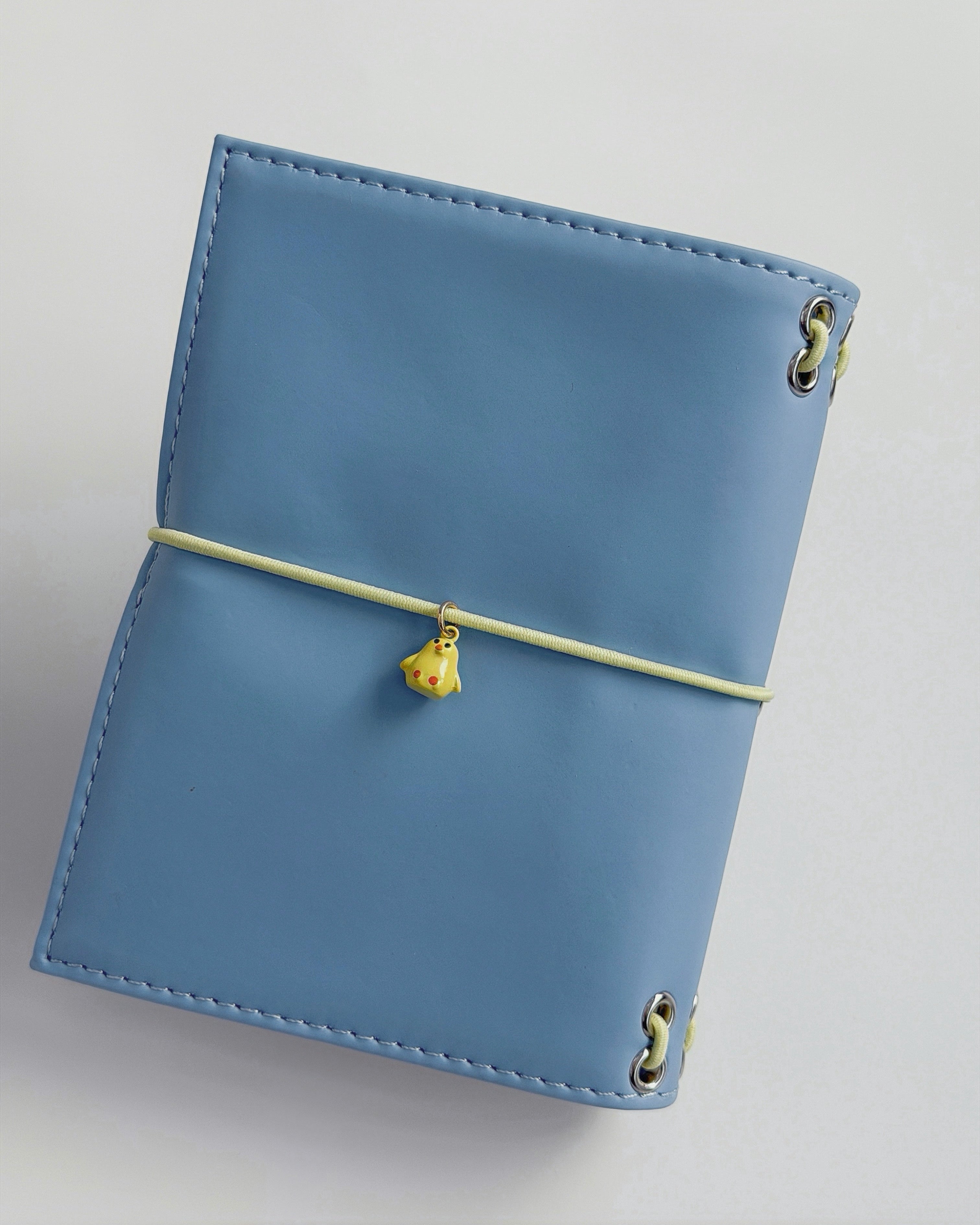 Back view of the Mist Blue pocket journal with a butter-yellow elastic band and yellow chick charm