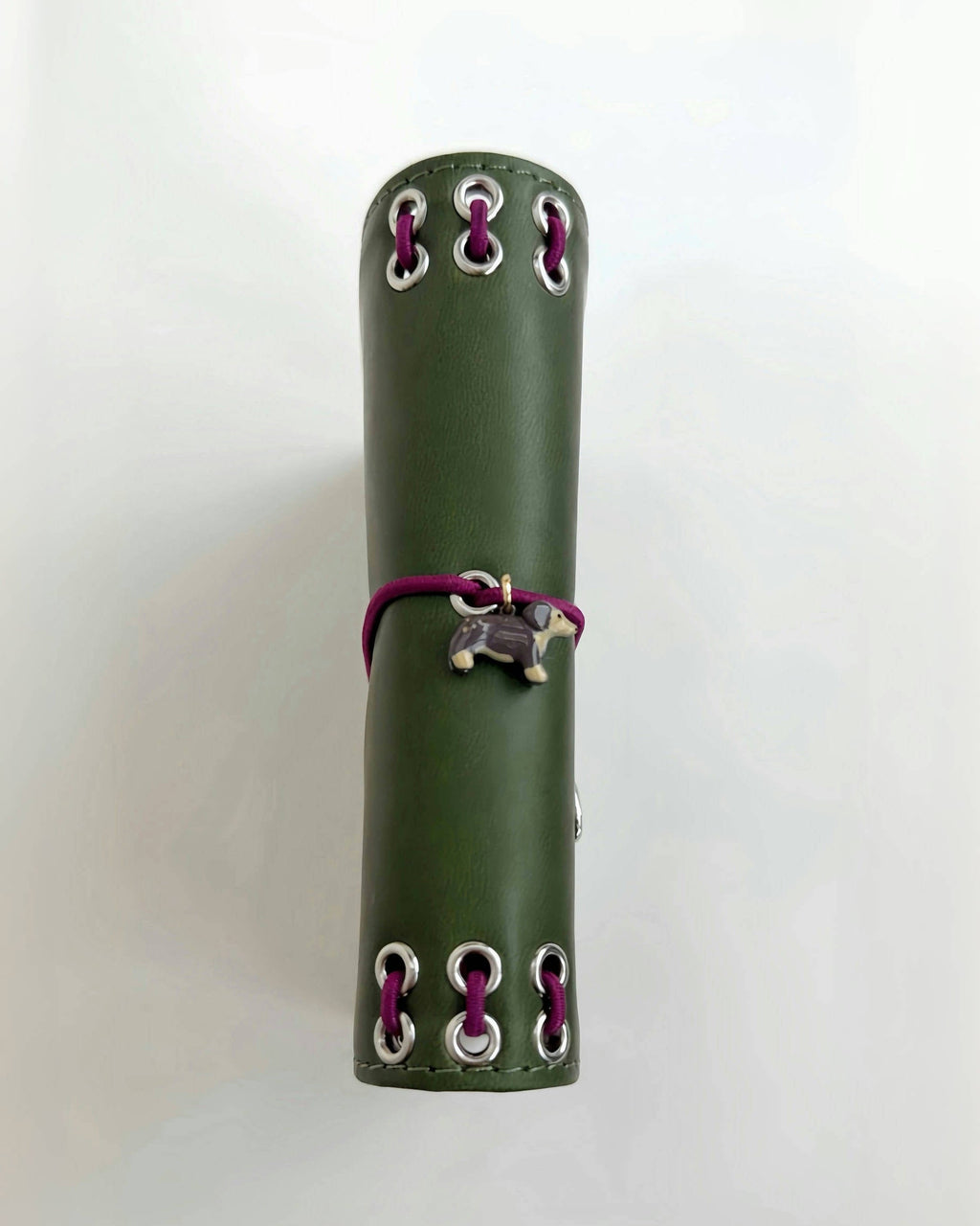 Side view of the Olive Pocket Journal showing its smooth PU cover, burgundy elastic band, dachshund charm, and metal eyelet binding on a white background.