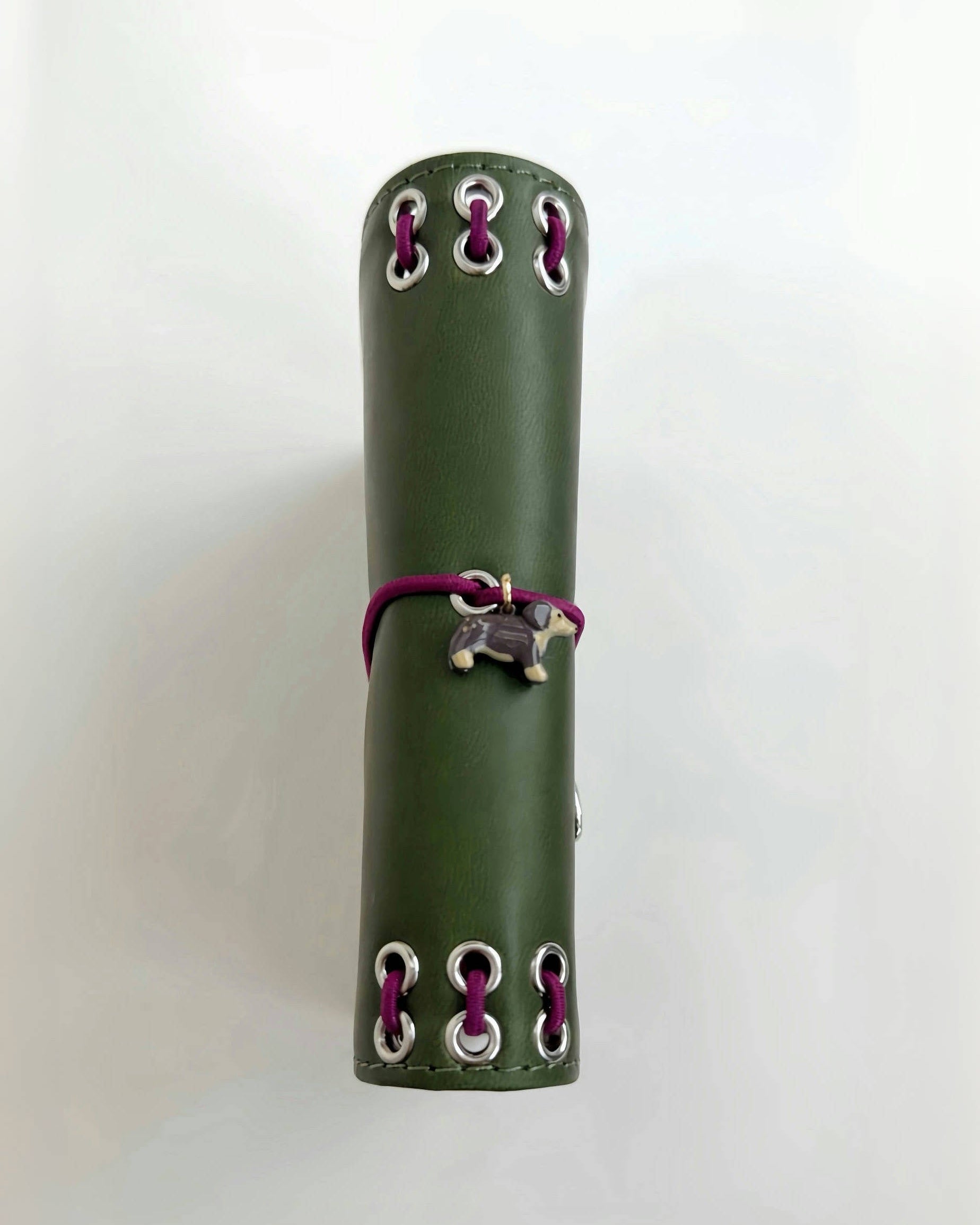 Side view of the Olive Pocket Journal showing its smooth PU cover, burgundy elastic band, dachshund charm, and metal eyelet binding on a white background.