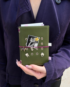 A person holds an Olive Pocket Journal styled with a burgundy elastic band, dachshund charm, four removable KEPT cover charms, and a black-and-white photo insert.