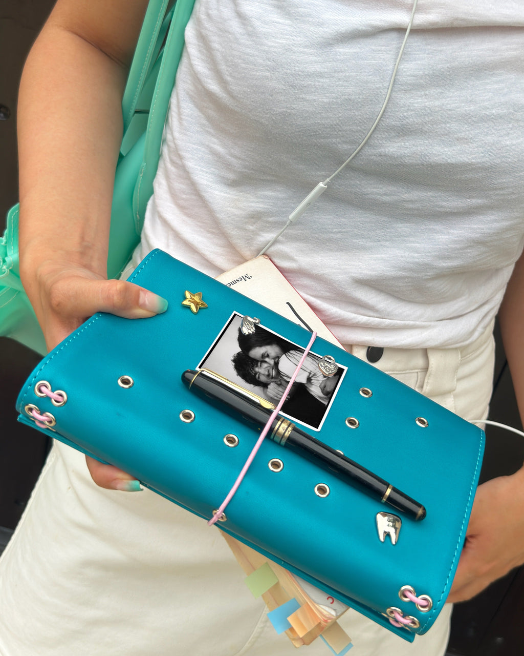 Person holding a teal full-size refillable journal styled with removable metal charms, a black-and-white photo, a pen, and pink elastic bands.
