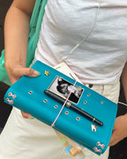 Person holding a teal full-size refillable journal styled with removable metal charms, a black-and-white photo, a pen, and pink elastic bands.