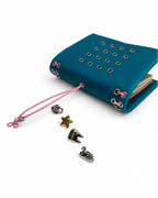 Teal pocket journal displayed at an angle with its blush-pink elastic band, scissors charm, and removable KEPT cover charms placed beside it