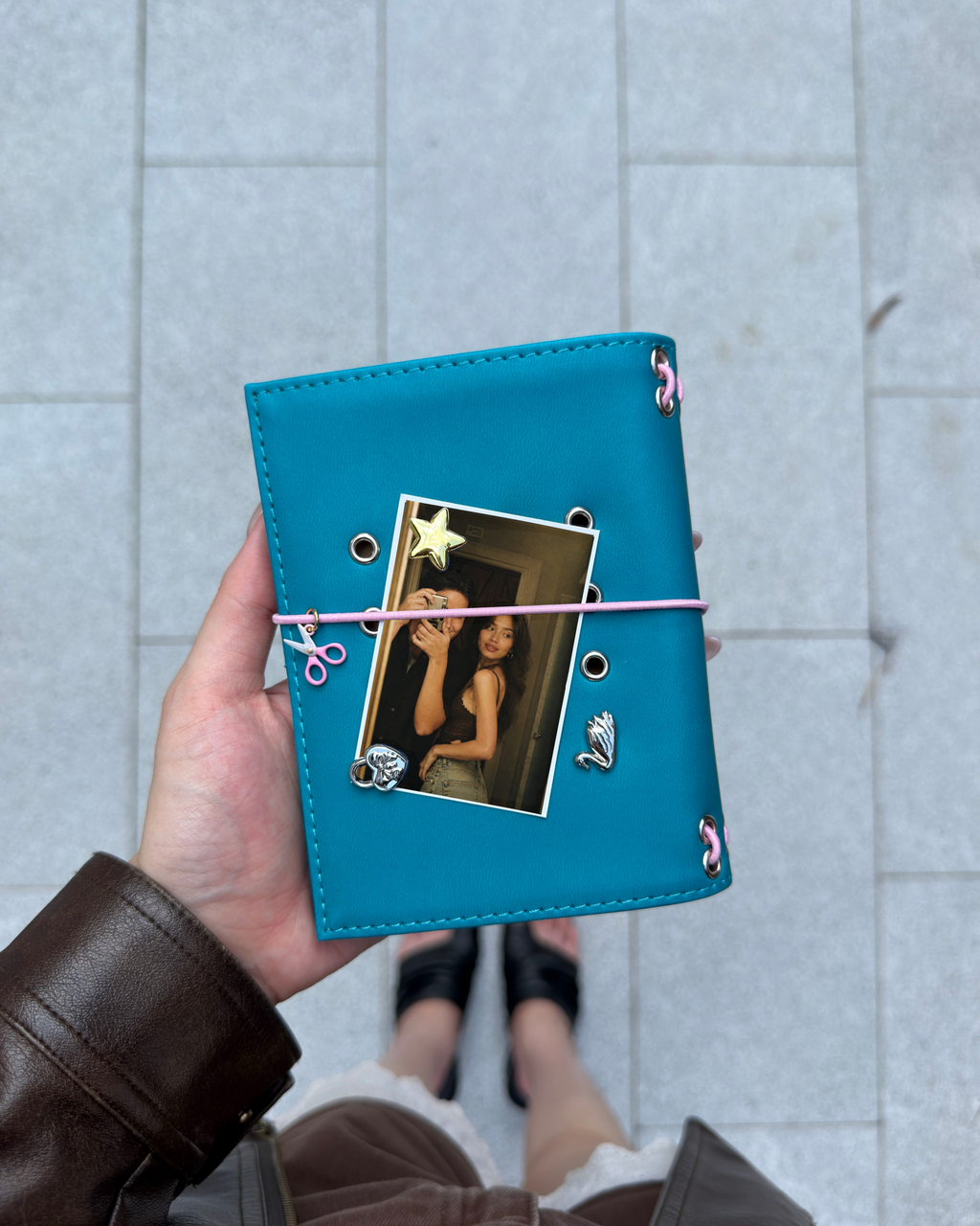 A hand holds a Teal Pocket Journal styled with a blush-pink elastic band, pink scissors charm, four removable KEPT cover charms, and a photo insert.